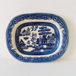 Op de afbeelding: Een antieke blauw-witte ovale serveerschaal. De schaal heeft een gedetailleerde landschapsafbeelding met pagodes, bomen en een brug. De rand heeft een decoratieve rand. Het keramische gerecht is een klassiek stuk vintage servies.