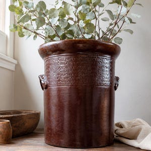 May include: A large, dark brown ceramic crock with a textured band around the middle and two small handles. The crock is filled with leafy green branches. The crock sits on a wooden surface.