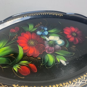 May include: Oval black serving tray with a hand-painted floral design. The tray features red, blue, white, and purple flowers with green leaves. The rim is decorated with a gold leaf pattern.
