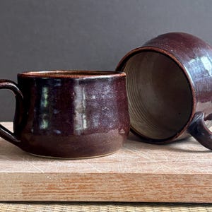Może przedstawiać: Dwa ceramiczne kubki z ciemną, błyszczącą glazurą w kolorze głębokiego fioletowego brązu. Jeden kubek stoi pionowo, ukazując swoją zewnętrzną stronę, podczas gdy drugi leży na boku, odsłaniając jasną, żebrowaną fakturę wnętrza oraz pasujące ciemne ucho.