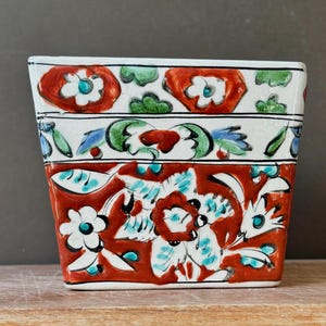 May include: A square ceramic planter featuring a hand-painted floral pattern in vibrant red, green, blue, and white. The design is divided into three horizontal bands with stylised blossoms, leaves, and intricate botanical motifs on a white base.