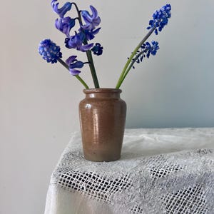 May include: A brown ceramic vase with a small bouquet of purple and blue hyacinths. The vase is sitting on a white crocheted tablecloth.