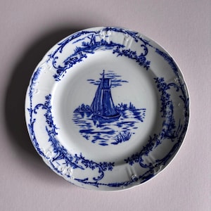 Puede incluir: Plato de cerámica blanca con una escena de velero azul en el centro. El plato tiene un borde festoneado y está decorado con patrones florales y paisajísticos azules alrededor del borde. El velero está sobre una masa de agua.