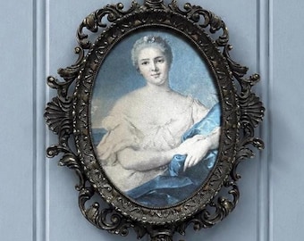 Puede incluir: Un retrato ovalado de una persona con cabello claro que viste una prenda blanca y un pañuelo azul, enmarcado en un marco metálico oscuro y ornamentado con volutas intrincadas y detalles florales en los bordes y los remates.