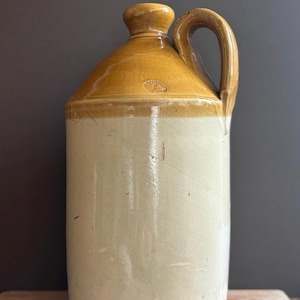 May include: A cylindrical stoneware jug featuring a two-tone glaze with a light beige body and a darker tan shoulder and neck. The jug has a single side handle, a narrow mouth, and a small circular stamp on the shoulder containing the number 5.