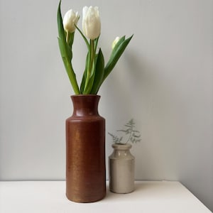 Könnte beinhalten: Eine braune Keramikvase mit weißen Tulpen und grünen Blättern. Daneben eine kleinere, hellfarbene Vase mit einem Zweig Grün. Die Vasen stehen auf einer weißen Oberfläche vor einer neutralen Wand.