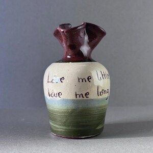 May include: A small, handmade ceramic vase with a ruffled rim. The vase features a color scheme of burgundy, cream, blue, and green. The words "Love me little, love me long" are handwritten on the cream-colored body of the vase.