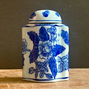 May include: A white ceramic jar with a blue floral pattern and a matching removable lid. The jar features a hexagonal shape with rounded corners and is decorated with blue painted flowers and leaves across the entire exterior surface.