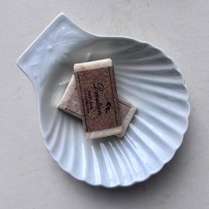 May include: A white, shell-shaped dish contains two wrapped soap bars. The soap wrappers are brown and feature the text "Dersat Sweet Soap". The dish has a ridged texture and a leaf design on one side.