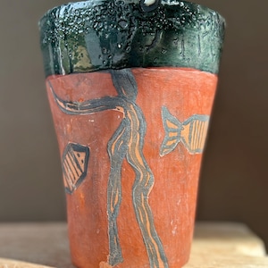May include: A terracotta-colored ceramic cup featuring a dark green, textured glaze around the rim. The body of the cup is decorated with etched, dark gray line drawings of a tall, slender figure and two fish, set against the matte orange surface.