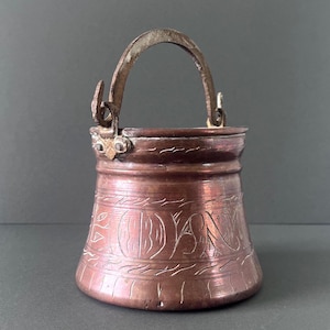 May include: A small, antique copper bucket with a curved handle. The bucket has etched designs and text around the body. The handle and the top rim are a darker, aged metal. The bucket is likely used for decorative purposes.