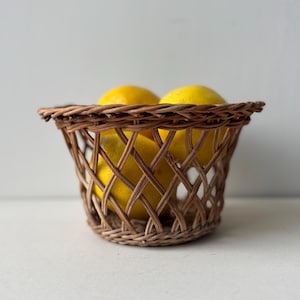 Op de afbeelding: Een bruine, geweven rieten mand gevuld met gele citroenen. De mand heeft een ronde vorm met een rasterpatroon. De citroenen zijn heldergeel en contrasteren met het bruin van de mand.