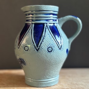 May include: A light blue ceramic pitcher with a textured, pebbled surface. The vessel features a dark blue glazed rim, a series of dark blue petal-shaped accents around the neck, and small dark blue circular patterns on the body with a handle.
