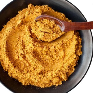 May include: A black bowl filled with a mound of yellow-brown spice powder. A wooden spoon is partially submerged in the powder, with some powder clinging to the spoon.