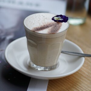 May include: A glass of creamy, light-colored beverage topped with a sprinkle of cinnamon and a single purple flower. The drink is served in a clear glass with a saucer.
