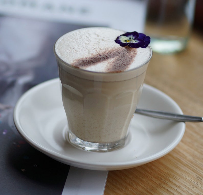 May include: A glass of creamy, light-colored beverage topped with a sprinkle of cinnamon and a single purple flower. The drink is served in a clear glass with a saucer.