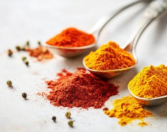 May include: A selection of vibrant spices, such as paprika and turmeric, displayed in silver spoons and scattered on a white surface. The image showcases a range of colours, including red, orange, and yellow. Green peppercorns are also visible.
