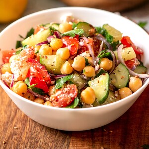 K&ouml;nnte beinhalten: Eine wei&szlig;e Sch&uuml;ssel gef&uuml;llt mit einem Salat aus Kichererbsen, Gurken, Tomaten, roten Zwiebeln und Petersilie. Der Salat ist mit Sesamsamen bestreut.