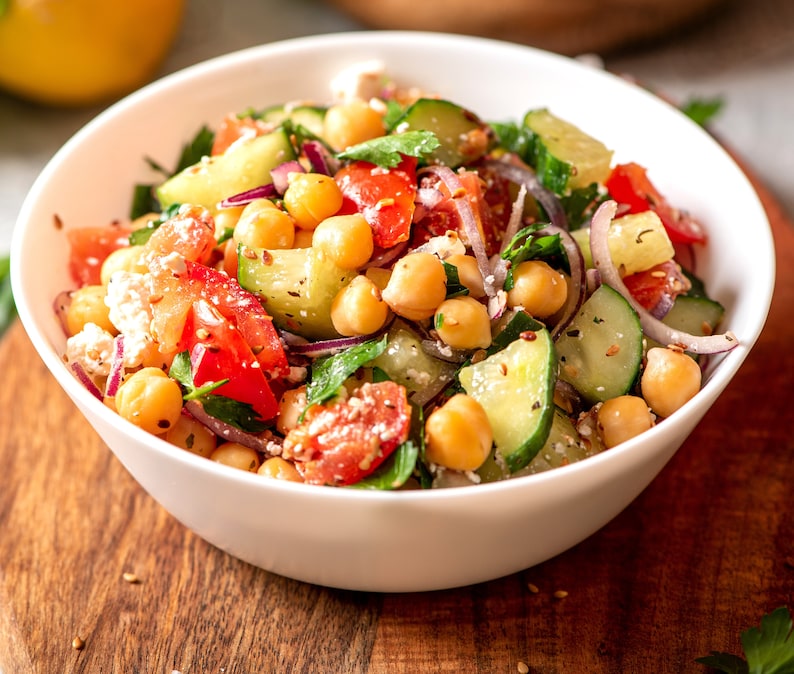 K&ouml;nnte beinhalten: Eine wei&szlig;e Sch&uuml;ssel gef&uuml;llt mit einem Salat aus Kichererbsen, Gurken, Tomaten, roten Zwiebeln und Petersilie. Der Salat ist mit Sesamsamen bestreut.