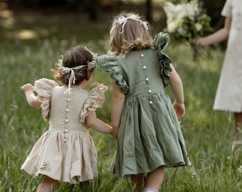 May include: Two young girls wearing white flower crowns and matching dresses walk through a field of green grass. The girl in the front is wearing a beige dress with a floral embroidery, while the girl in the back is wearing a sage green dress with ruffled sleeves.