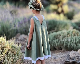 May include: A young person wearing a sleeveless olive green dress with white trim and white flats, standing outdoors. The dress has a simple, flowing design. A floral crown adorns the person's hair. The background features a natural setting with greenery.