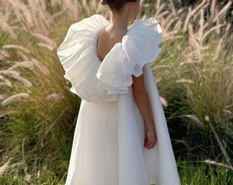 May include: A young girl wearing a white dress with a large ruffled collar. The dress has a flowing skirt and is made of a lightweight fabric. The girl is standing in a field of tall grass.