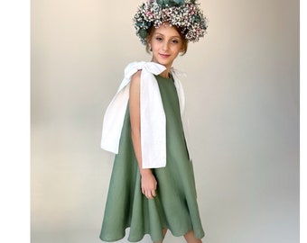 May include: A young person wearing a green linen dress with white bow details and a flower crown. The dress has a flowy skirt and the person is wearing white shoes.