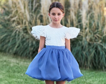 Può includere: Una giovane persona indossa una camicetta bianca di pizzo con maniche a sbuffo e una gonna di tulle blu. La gonna è raccolta in vita e ha una forma gonfia e voluminosa. La persona è in piedi in un'area erbosa con alte piante piumate sullo sfondo.