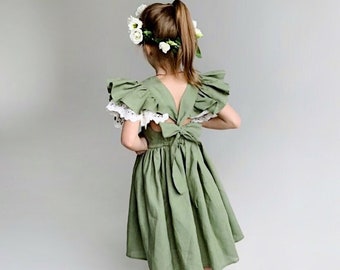 May include: A little girl wearing a sage green dress with a white lace trim and a white flower crown. The dress has a ruffled neckline and a bow in the back.