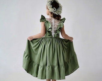 May include: A young girl wearing a sage green dress with a white lace back and a flower crown. The dress has a tiered skirt and ruffled sleeves.