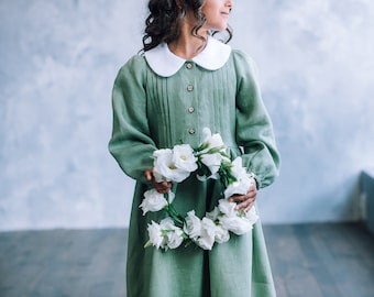Op de afbeelding: Een saliegroene jurk met een witte kraag en knopen. De jurk wordt gecombineerd met een witte bloemenkrans. De jurk heeft lange mouwen en een geplooid lijfje. Het algemene thema is een vintage of bohemien stijl.