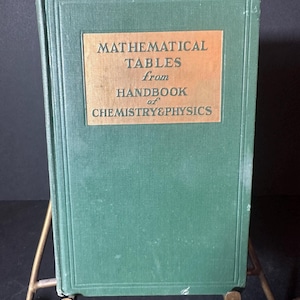 May include: A vintage green book titled "MATHEMATICAL TABLES from HANDBOOK of CHEMISTRY & PHYSICS" with a gold label. The book is displayed on a brass easel, showcasing its cover.
