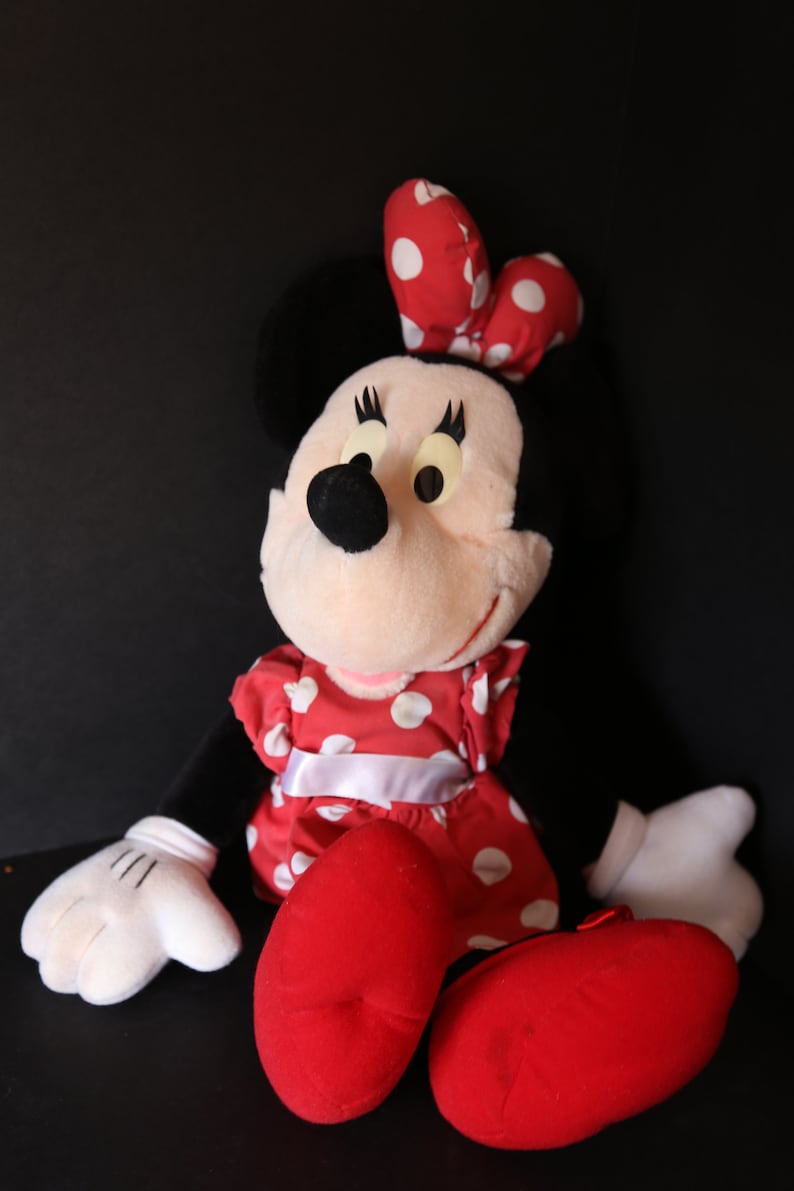 Pu&ograve; includere: Un giocattolo di peluche di Minnie Mouse che indossa un vestito rosso con pois bianchi e un fiocco rosso con pois bianchi. Ha le orecchie nere e un naso nero. &Egrave; seduta con le gambe incrociate e le mani in grembo.