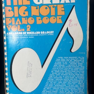 Puede incluir: Un libro de piano vintage titulado "The Great Big Note Piano Book Vol. 2" con una cubierta azul y letras naranjas. La portada presenta una gran nota musical y una lista de canciones. El libro está arreglado por Richard Bradley.