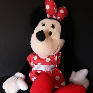 Pu&ograve; includere: Un giocattolo di peluche di Minnie Mouse che indossa un vestito rosso con pois bianchi e un fiocco rosso con pois bianchi. Ha gli occhi neri, un naso nero e guanti bianchi. &Egrave; seduta con le gambe incrociate e i suoi piedi sono rossi.
