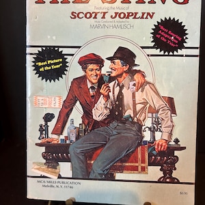 May include: A vintage book cover for "The Sting" movie, featuring two men in period attire. The cover includes text such as "Winner of 7 Academy Awards" and "Best Picture of the Year". The artwork has a retro style with a light blue background.