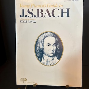Puede incluir: Un libro antiguo titulado "Young Pianist's Guide to J.S. Bach" con un retrato de J.S. Bach. La portada es de color crema con texto dorado y azul oscuro. El libro está apoyado en un soporte de metal decorativo.