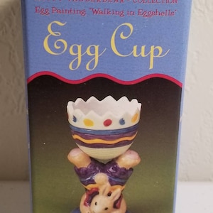 May include: A ceramic egg cup shaped like a bunny holding up a colorful egg. The egg cup is decorated with purple, yellow, and white stripes and dots. The bunny is wearing purple pants and has a red bow tie. The box reads "Muffy Vanderbear® Collection Egg Painting "Walking in Eggshells" Egg Cup Hoppy™".