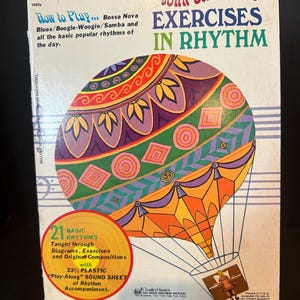 May include: Vintage music book titled "John Brimhall's Exercises in Rhythm." The cover features a colorful hot air balloon design with text that reads "How to Play... Bossa Nova, Blues/Boogie-Woogie/Samba." Includes a "Play-Along" sound sheet.