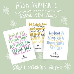 May include: Three white cards with quotes from musicals, including "Wicked," "Hamilton," and "Mamma Mia," are displayed against a light green background with snowflakes. The cards feature colorful text and are labeled "Brand New Prints!" and "Great Stocking Fillers!"