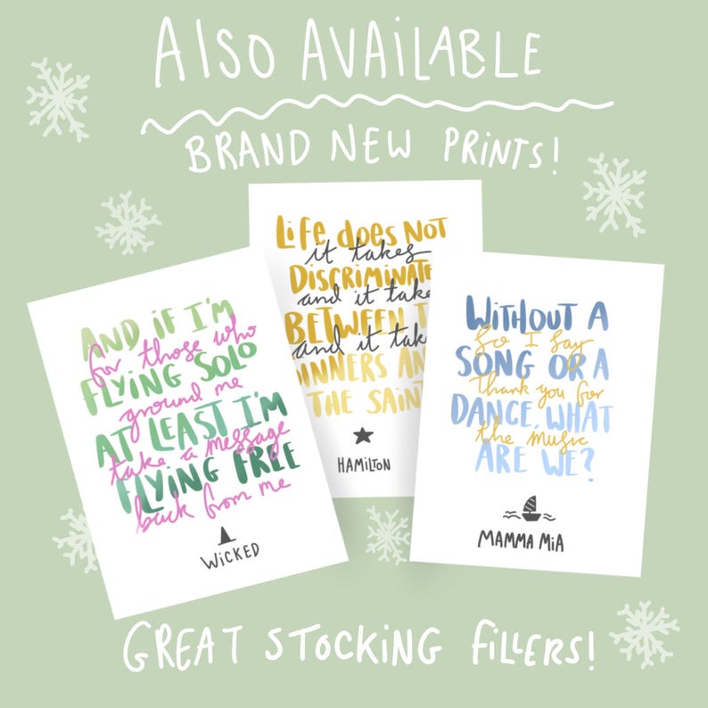 May include: Three white cards with quotes from musicals, including "Wicked," "Hamilton," and "Mamma Mia," are displayed against a light green background with snowflakes. The cards feature colorful text and are labeled "Brand New Prints!" and "Great Stocking Fillers!"
