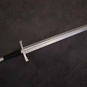 Puede incluir: Una espada plateada con una guarda en forma de cruz y un mango negro. La hoja es recta y termina en punta. El pomo de la espada es circular. El diseño evoca una estética medieval o fantástica. La longitud de la espada es de aproximadamente 76 cm.