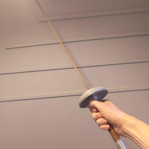 May include: A fencing foil, held in a hand, against a neutral background. The foil has a long, thin, flexible blade, a circular hand guard, and a grip. The grip is wrapped in a brown material, and the pommel is silver.