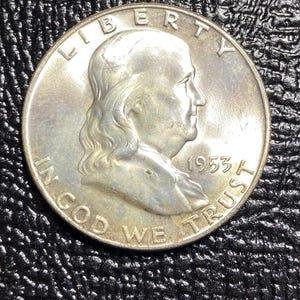 May include: A silver coin featuring a profile of a man, with the words "LIBERTY" and "IN GOD WE TRUST" inscribed. The year "1953" is also visible. The coin is set against a textured black background.