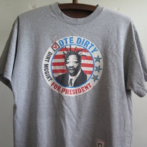 Pu&ograve; includere: Maglietta grigia con una grafica di un uomo con i dreadlocks e uno slogan di campagna presidenziale. Lo slogan recita "Vote Dirty Dirt McGirt for President".