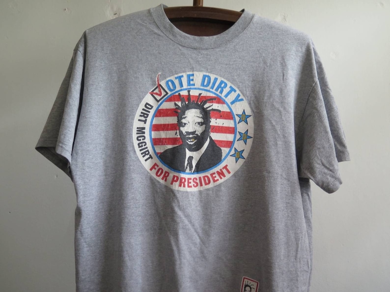 Pu&ograve; includere: Maglietta grigia con una grafica di un uomo con i dreadlocks e uno slogan di campagna presidenziale. Lo slogan recita "Vote Dirty Dirt McGirt for President".