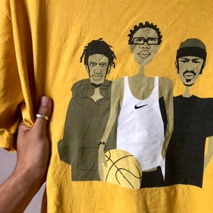 May include: Yellow t-shirt with a graphic print of three cartoon men. The men are wearing different outfits, including a white tank top with a Nike logo, a green hooded sweatshirt, and a black t-shirt. The men are holding a basketball.