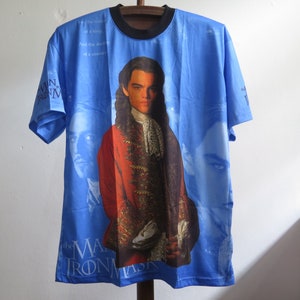 May include: Blue t-shirt featuring a portrait of a man in a red coat with the text "The Man in the Iron Mask." The shirt has a black collar and short sleeves. The background is a light blue color with additional portraits.