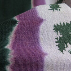 May include: Close-up of a tie-dye fabric with a purple and green colour scheme. The fabric has a white background with green and purple dye creating a swirling pattern. The fabric has a textured surface.