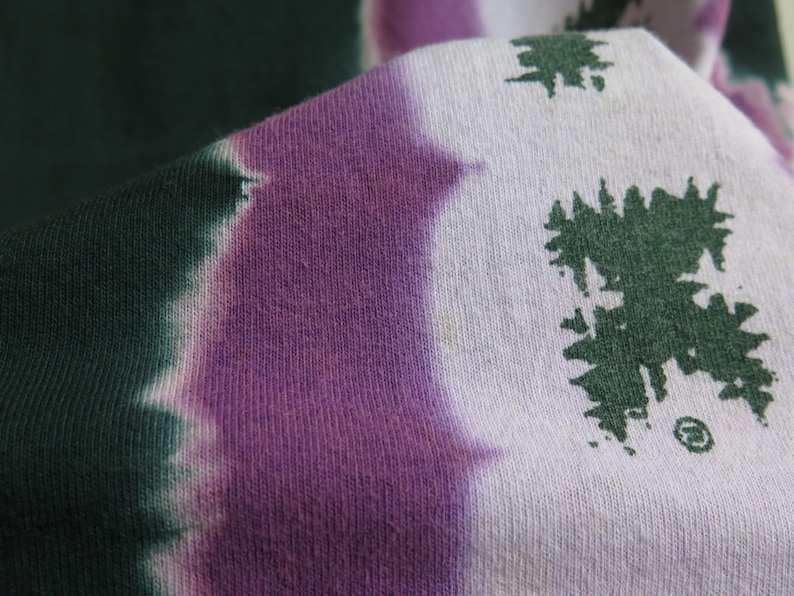 May include: Close-up of a tie-dye fabric with a purple and green colour scheme. The fabric has a white background with green and purple dye creating a swirling pattern. The fabric has a textured surface.